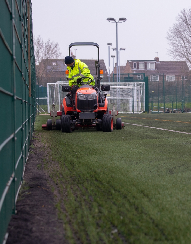 man on mower brushing an artificial pitch carpet next to fence